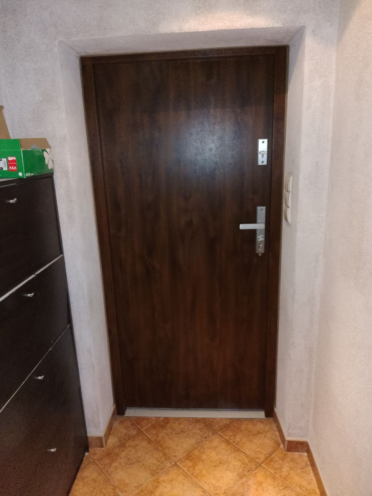 Brązowe drzwi wewnętrzne z metalową klamką i zamkiem, widoczne na tle beżowej ściany i brązowej podłogi z płytek, obok czarna szafka z pudełkiem na górze.