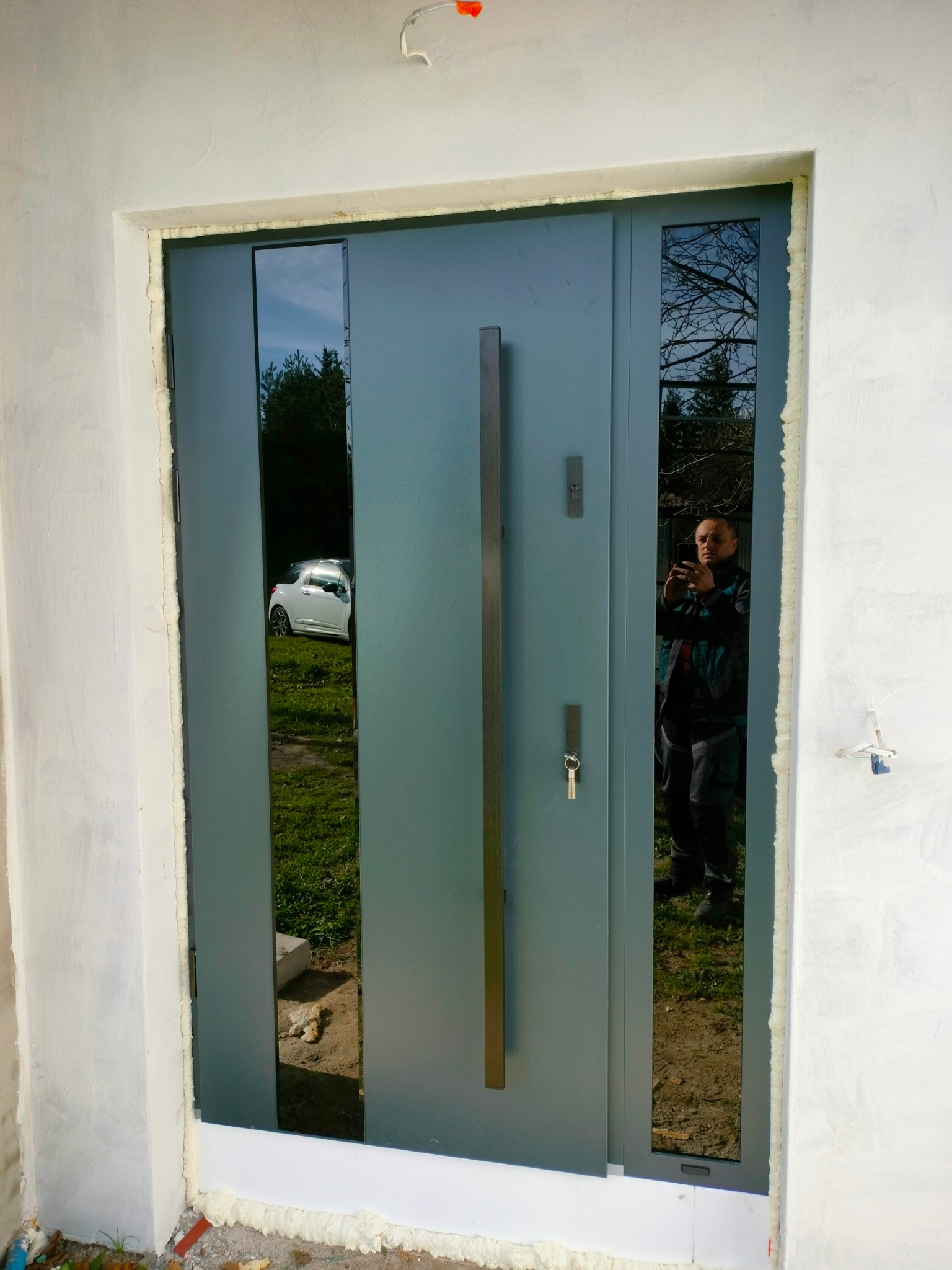 Szare drzwi wejściowe z długim uchwytem i szybami bocznymi, widoczny montaż z pianką uszczelniającą, odbicie osoby robiącej zdjęcie w szybie.