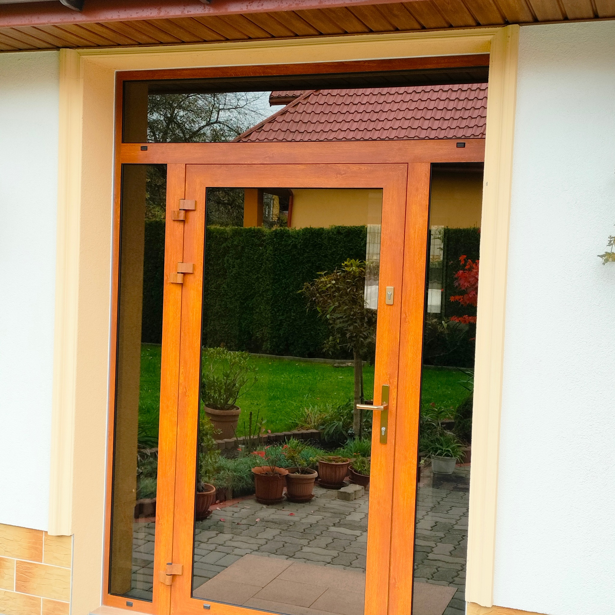 Drewniane drzwi zewnętrzne z szybą, widok na ogród z kostką brukową i roślinami w donicach, jasna elewacja budynku.
