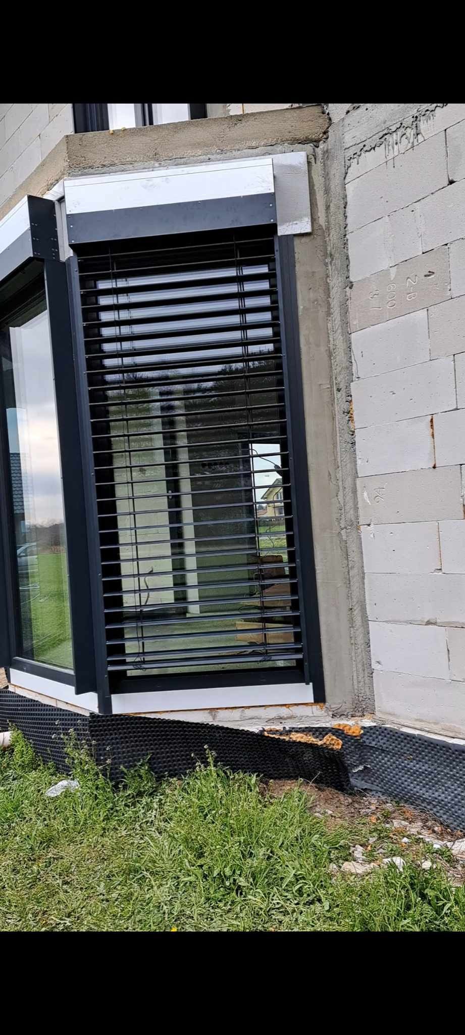 Ciemne żaluzje poziome zamontowane na oknie budynku w trakcie budowy, widoczny surowy mur i fragment trawnika.