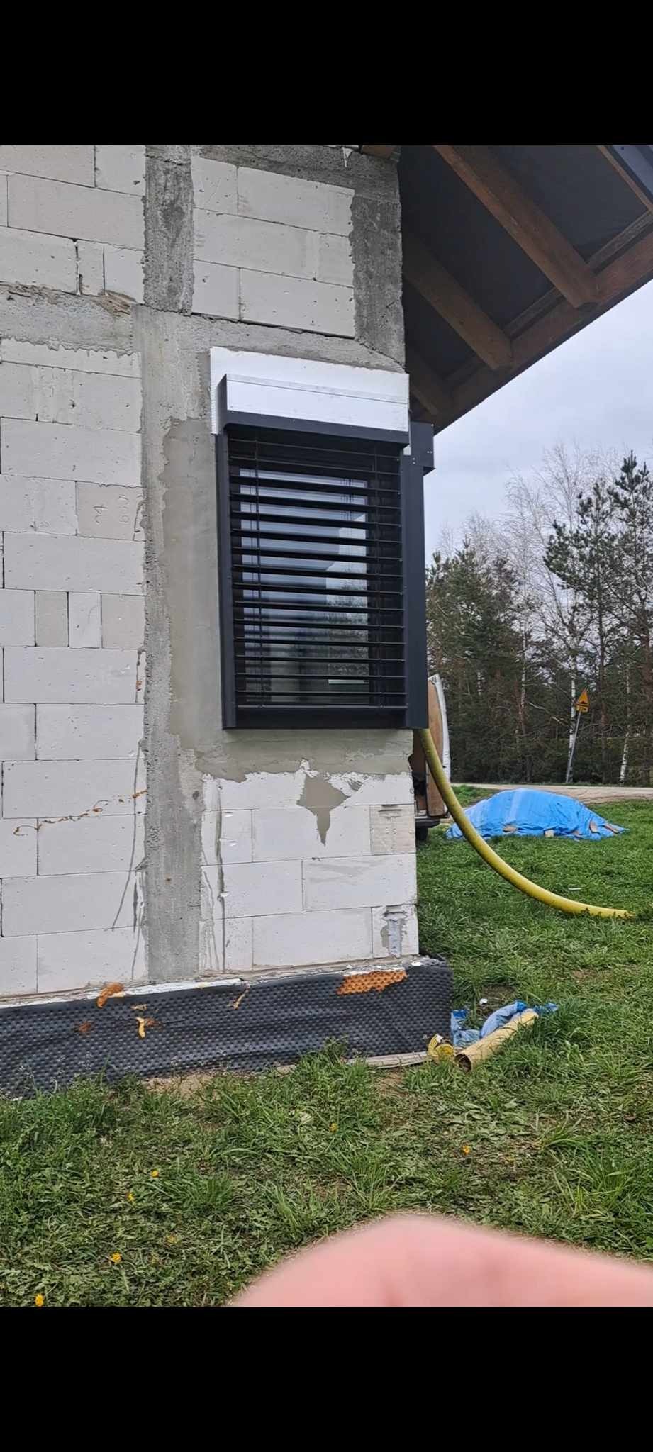Zamontowana czarna roleta zewnętrzna na oknie w surowym murze z bloczków, widoczny fragment dachu, żółty wąż i niebieska plandeka na trawie.
