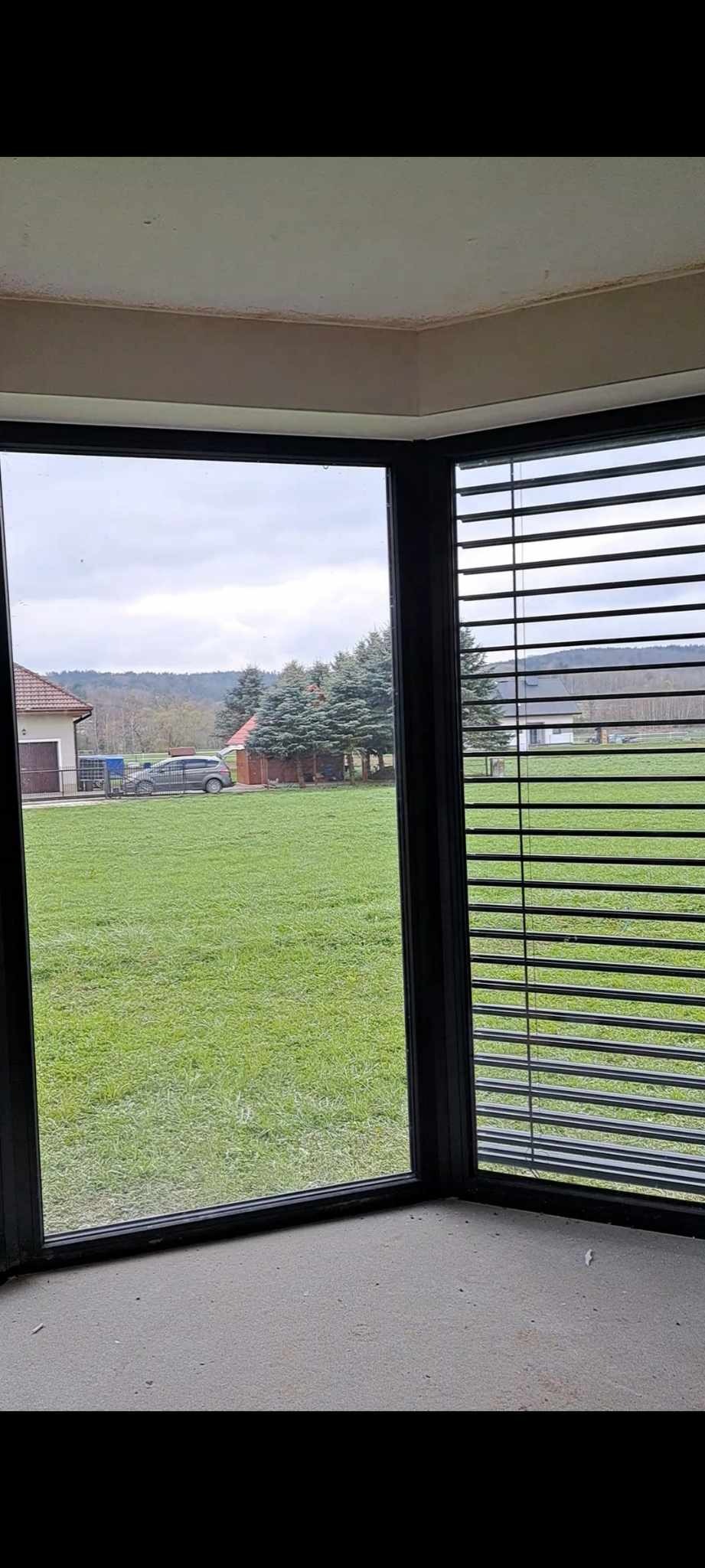Wnętrze pokoju z narożnym oknem PCV z czarną ramą i zamontowanymi poziomymi żaluzjami, widok na trawnik, dom i zaparkowany samochód w pochmurny dzień.