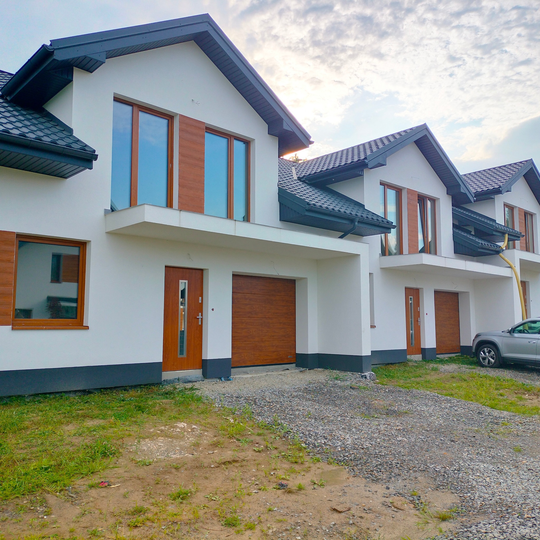 Szereg nowych domów jednorodzinnych z drewnopodobnymi bramami garażowymi i oknami, widok z zewnątrz, częściowo zagospodarowany teren.