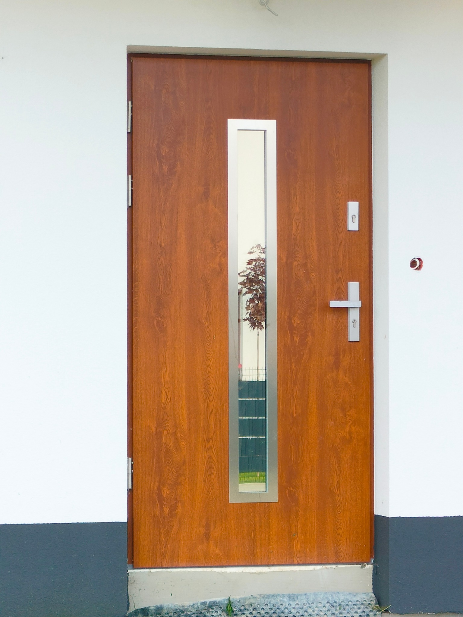 Brązowe drzwi wejściowe z pionowym, srebrnym przeszkleniem odbijającym drzewo, zamontowane w białej ścianie z szarą podmurówką.