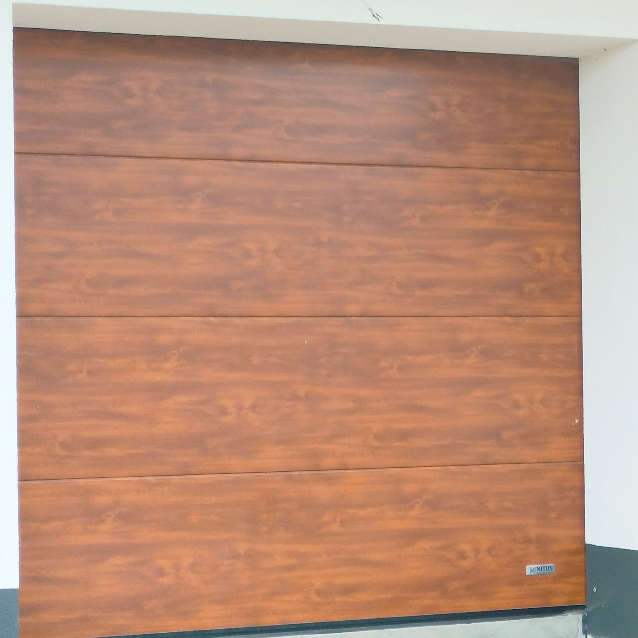 Zamknięta segmentowa brama garażowa w kolorze drewna, z widocznym logo producenta Nitus w prawym dolnym rogu, zamontowana w jasnym garażu.