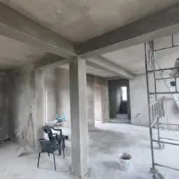 Wnętrze w stanie deweloperskim z surowymi ścianami i sufitem po tynkowaniu, widoczne rusztowanie, krzesła i butelki wody, ślady po pracach wykończeniowych.