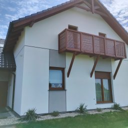 GROBUD KRZYSZTOF GROBAREK - Nowoczesny dom jednorodzinny z białymi ścianami, drewnianym balkonem i brązową stolarką okienną, otoczony zielenią i kamieniami ozdobnymi.