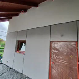 Elewacja budynku po tynkowaniu i malowaniu z widocznymi panelami, oknem zabezpieczonym folią i zakrytymi drzwiami, na dole rozłożona folia ochronna.