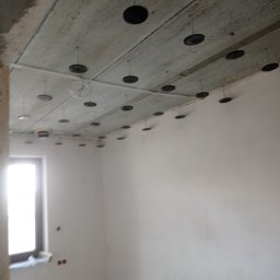GROBUD KRZYSZTOF GROBAREK - Surowy betonowy sufit z zamocowanymi czarnymi dyskami izolacyjnymi i metalowymi prętami, przygotowany do dalszych prac wykończeniowych w jasnym pomieszczeniu z oknem.