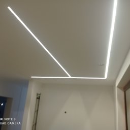 GROBUD KRZYSZTOF GROBAREK - Nowoczesne oświetlenie sufitowe z liniowymi lampami LED tworzącymi geometryczny wzór w świeżo wykończonym wnętrzu z drabiną malarską w rogu.
