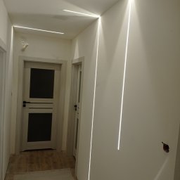 GROBUD KRZYSZTOF GROBAREK - Nowoczesny korytarz z liniowym oświetleniem LED wpuszczonym w ściany i sufit, widoczne dwie drewniane drzwi, podłoga w trakcie wykończenia, surowe ściany przygotowane do malowania.