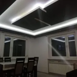 Nowoczesny salon z czarno-białym sufitem podwieszanym z oświetleniem LED, stół z krzesłami, okna i telewizor na ścianie.