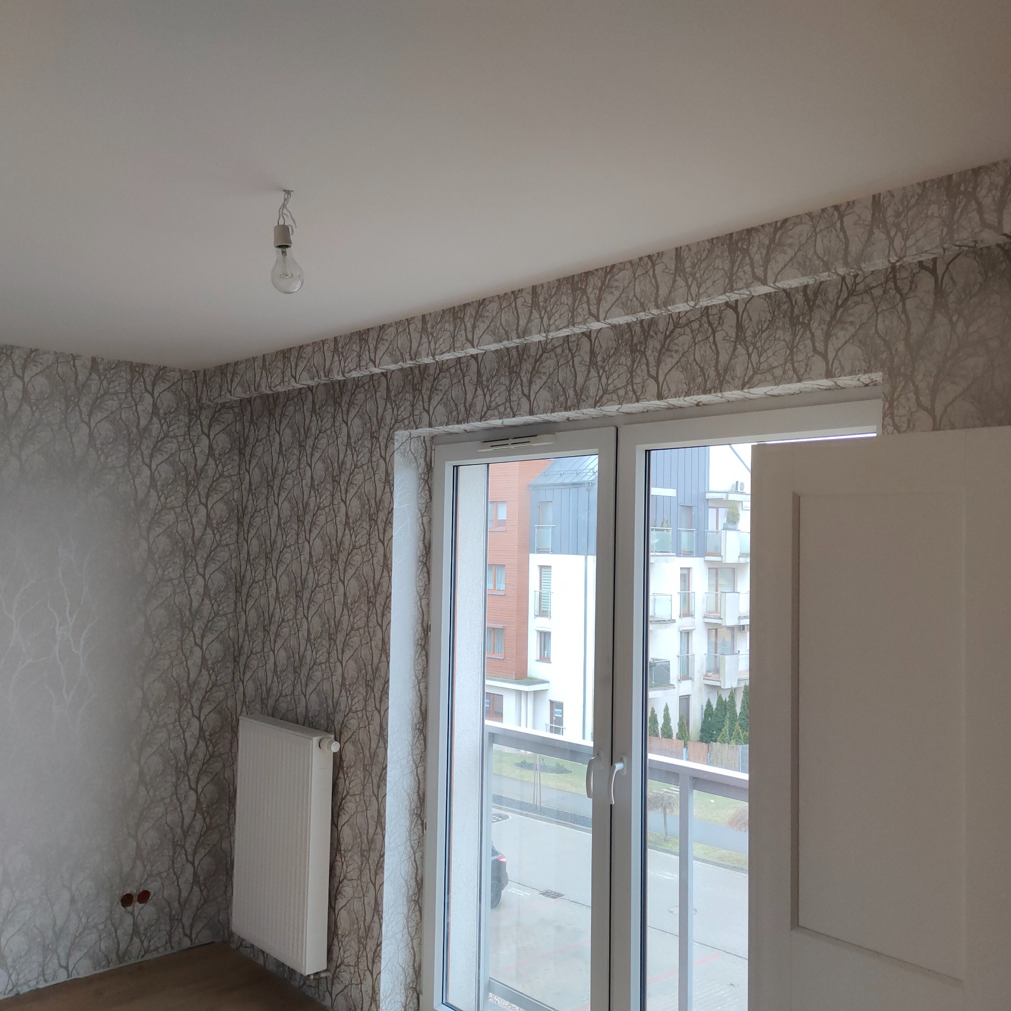 Pokój po remoncie z tapetą w motywie drzew, biały grzejnik i okno balkonowe z widokiem na budynek mieszkalny. Na suficie zwisa żarówka na kablu.