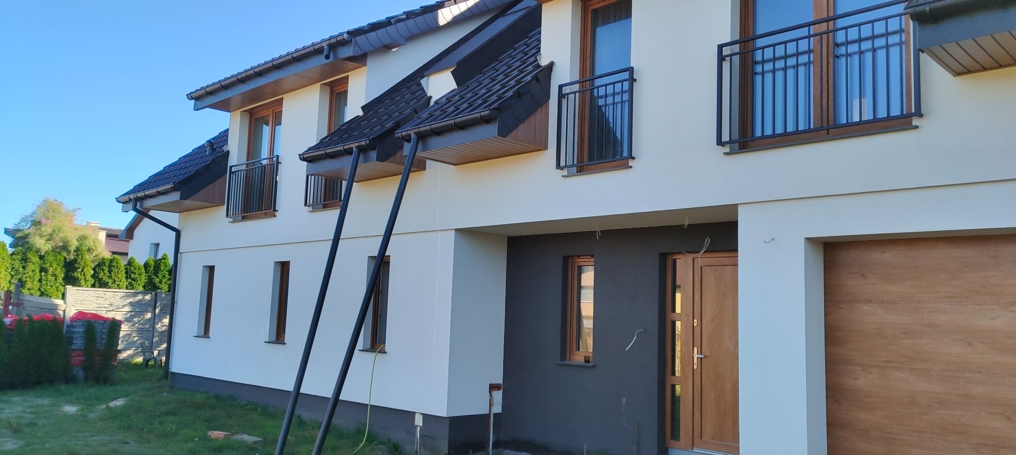 Nowo wykończona elewacja dwukondygnacyjnego domu z balkonami i rynnami spustowymi, fragment ściany pomalowany na szaro, widoczne przewody elektryczne wystające ze ściany.