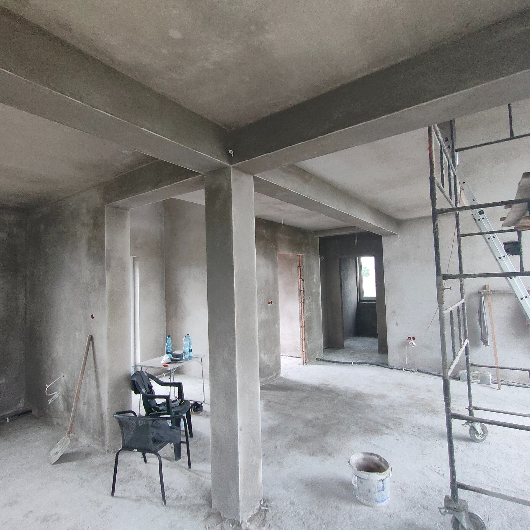 Wnętrze w stanie deweloperskim z surowymi ścianami i sufitem po tynkowaniu, widoczne rusztowanie, krzesła i butelki wody, ślady po pracach wykończeniowych.