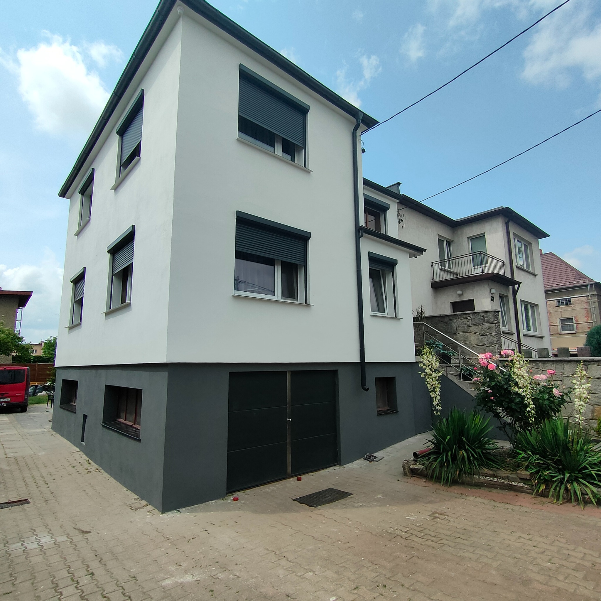 Dwukondygnacyjny dom po odświeżeniu elewacji, z szarym cokołem i białymi ścianami, okna z roletami zewnętrznymi, widok z poziomu chodnika.