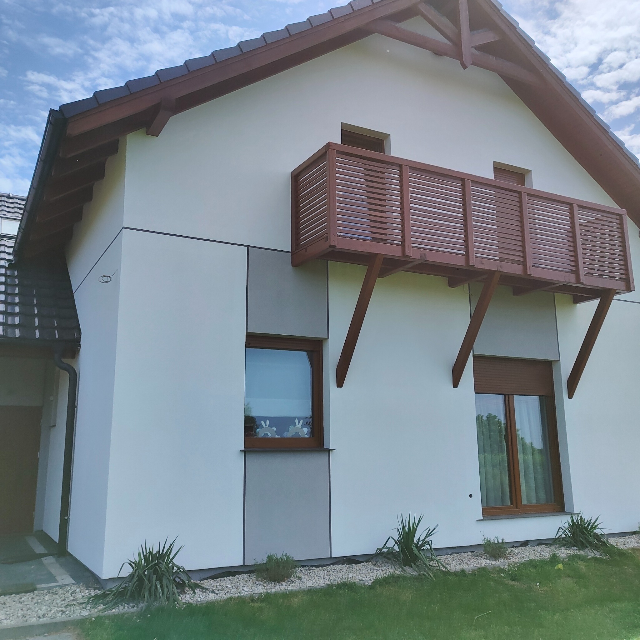 Nowoczesny dom jednorodzinny z białymi ścianami, drewnianym balkonem i brązową stolarką okienną, otoczony zielenią i kamieniami ozdobnymi.
