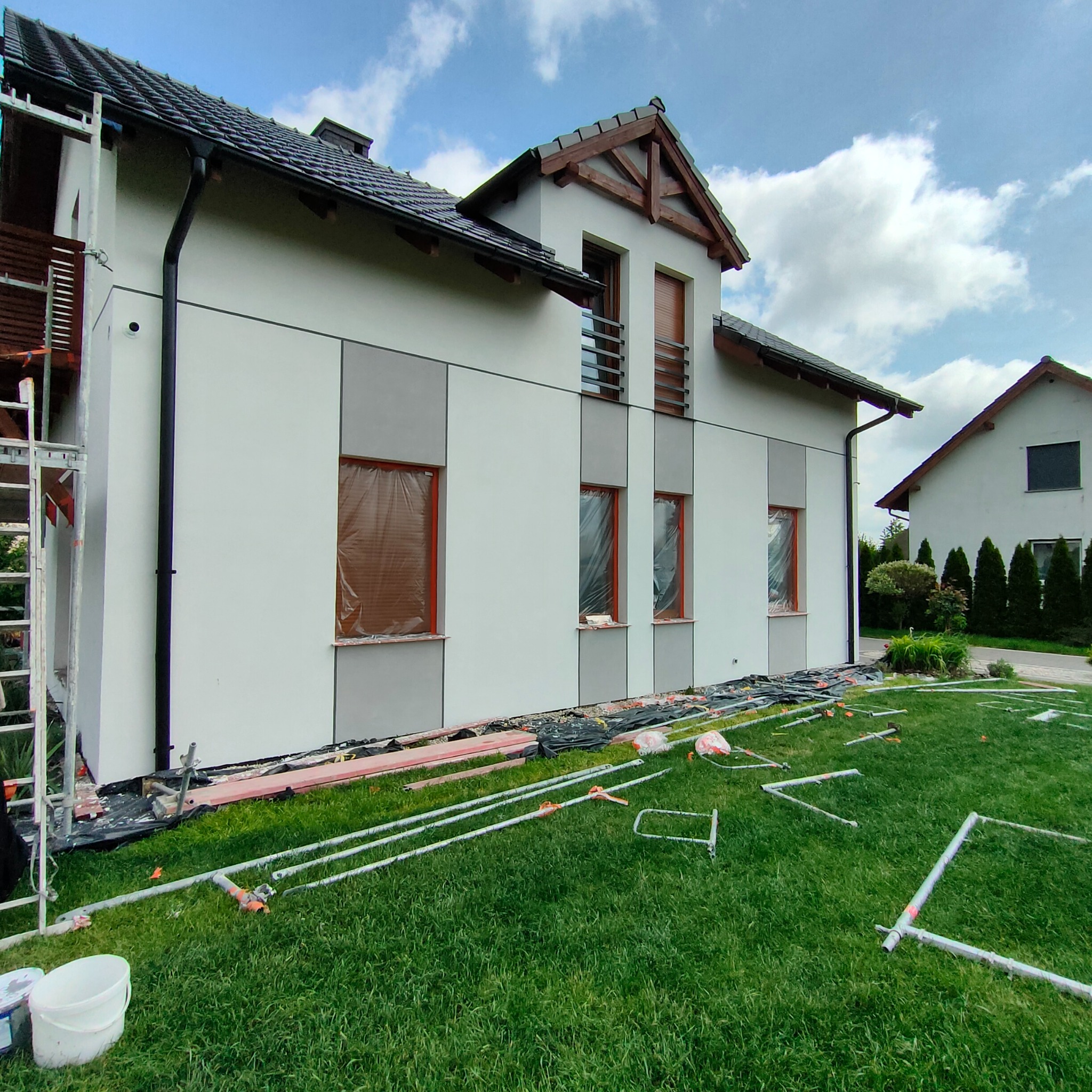 Dwukondygnacyjny dom jednorodzinny z szarą elewacją w trakcie malowania, okna zabezpieczone folią, na trawniku rozłożone elementy rusztowania i folie ochronne.