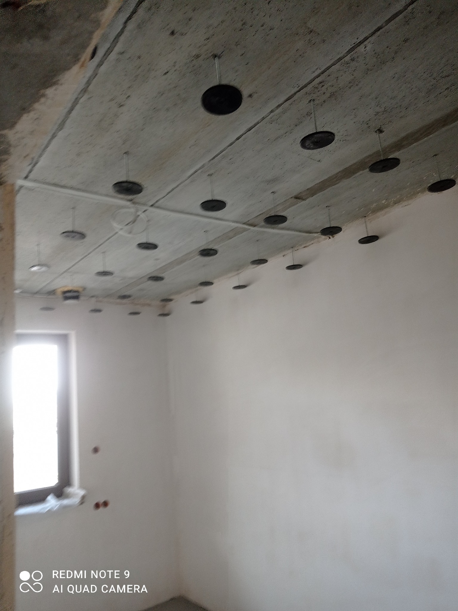 Surowy betonowy sufit z zamocowanymi czarnymi dyskami izolacyjnymi i metalowymi prętami, przygotowany do dalszych prac wykończeniowych w jasnym pomieszczeniu z oknem.
