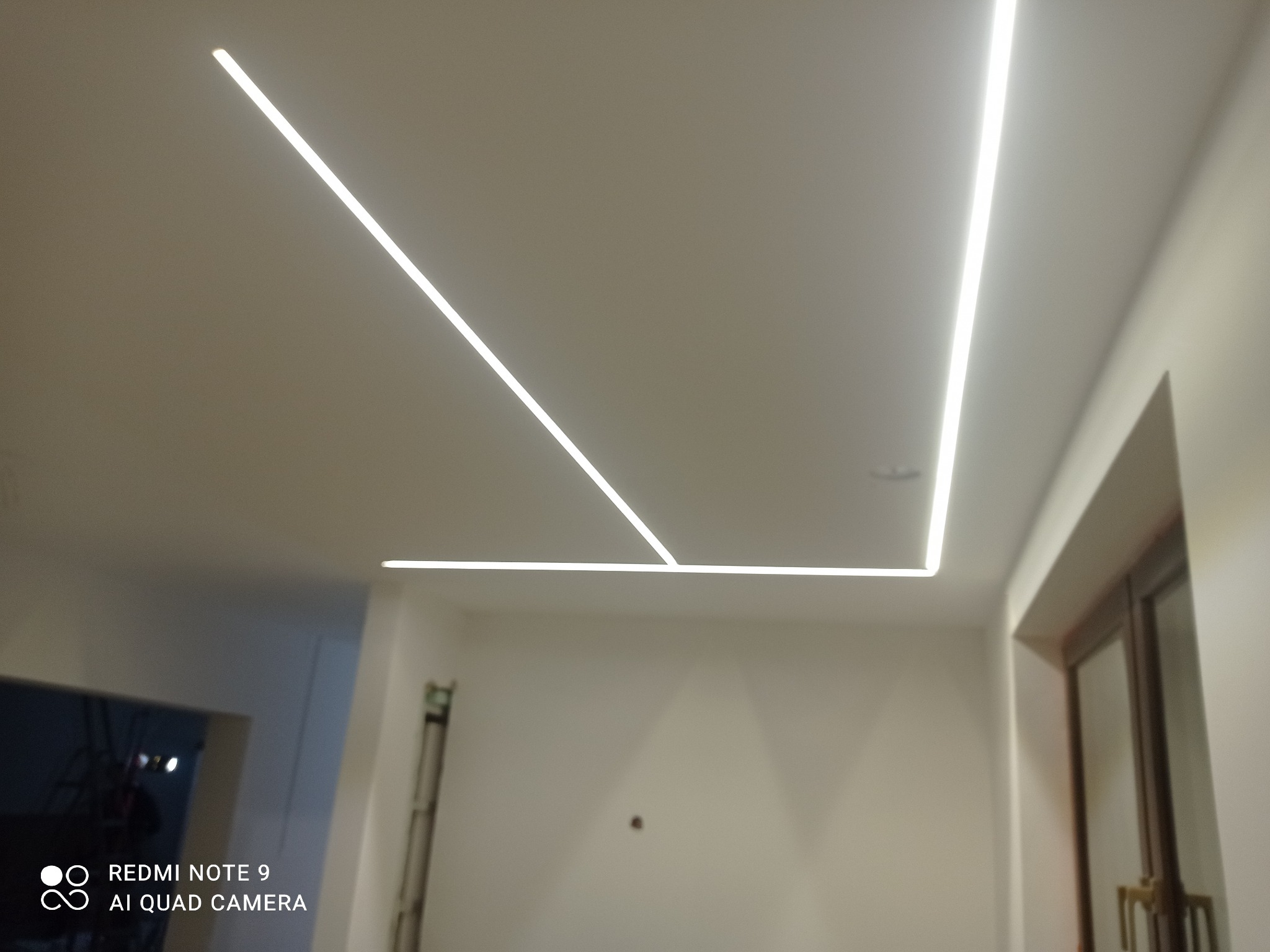 Nowoczesne oświetlenie sufitowe z liniowymi lampami LED tworzącymi geometryczny wzór w świeżo wykończonym wnętrzu z drabiną malarską w rogu.