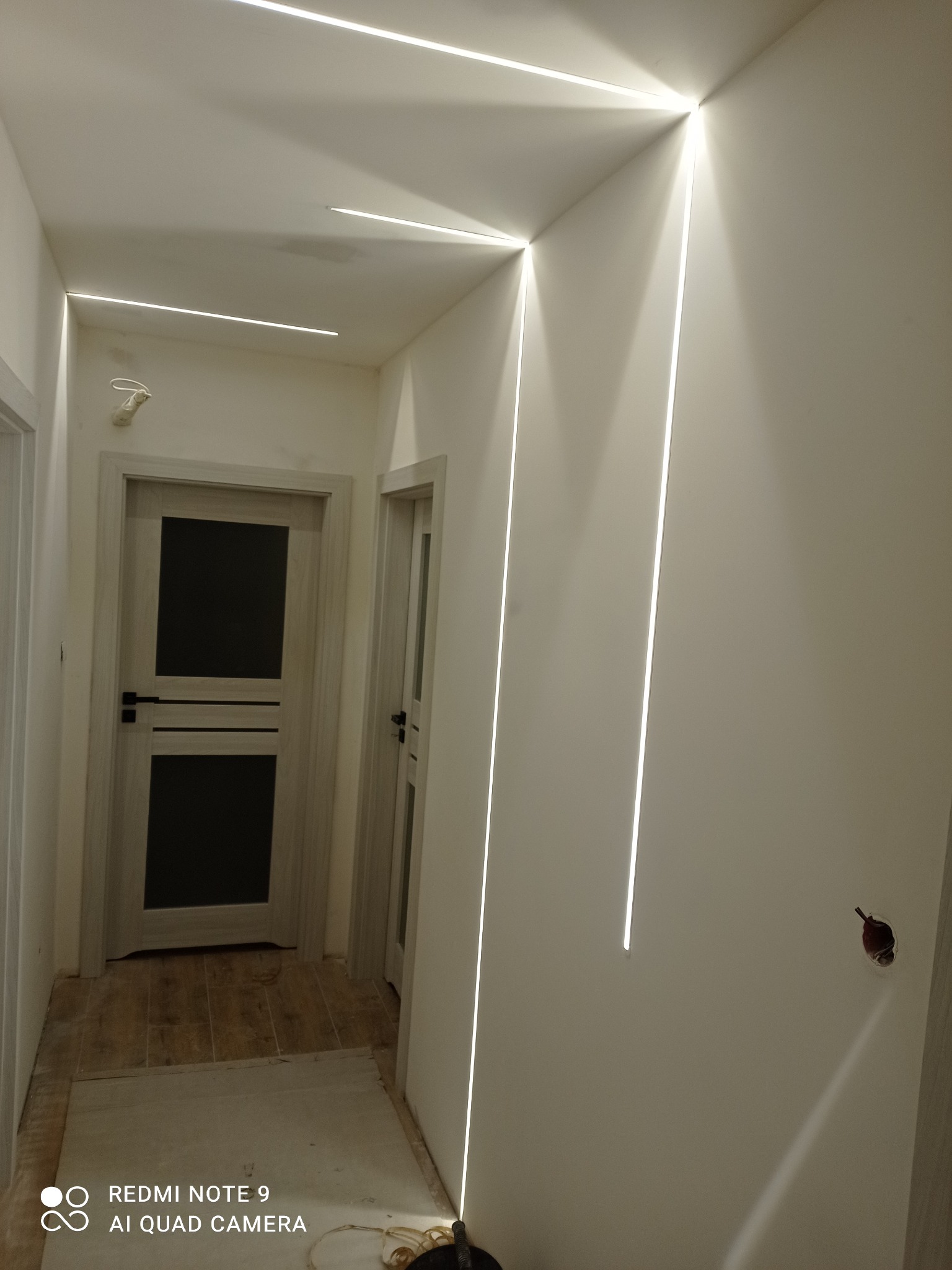 Nowoczesny korytarz z liniowym oświetleniem LED wpuszczonym w ściany i sufit, widoczne dwie drewniane drzwi, podłoga w trakcie wykończenia, surowe ściany przygotowane do malowania.