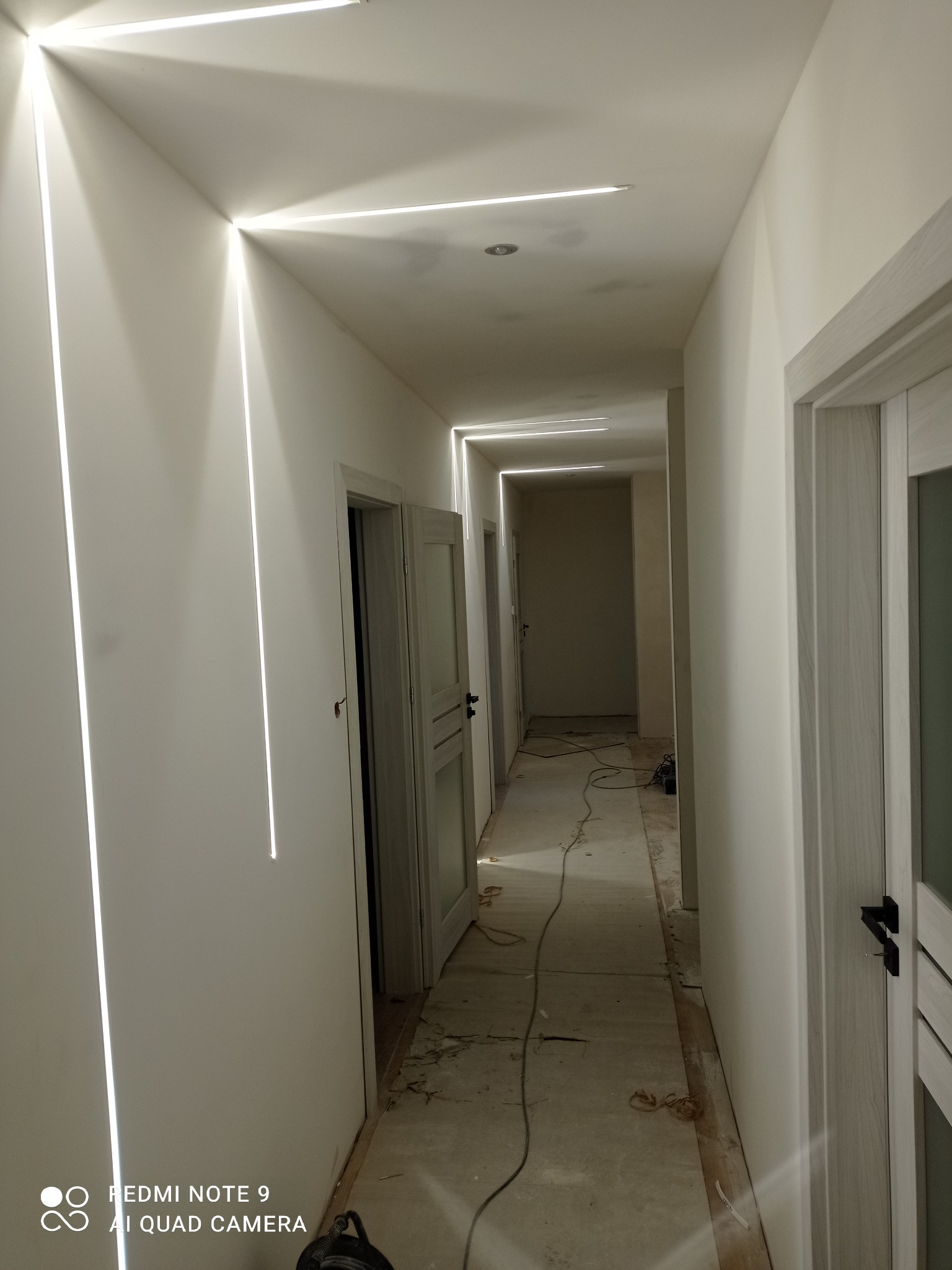 Korytarz w trakcie wykańczania w Ostrowie Wielkopolskim, z minimalistycznym oświetleniem liniowym wpuszczonym w sufit i ściany, widoczne otwarte drzwi i przygotowane podłoże.