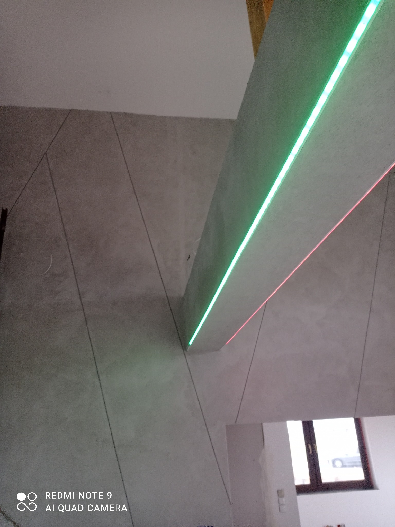 Ściana wykończona gładzią z dekoracyjnymi liniami i podświetleniem LED w kolorach zielonym i czerwonym, widoczny fragment okna w tle.