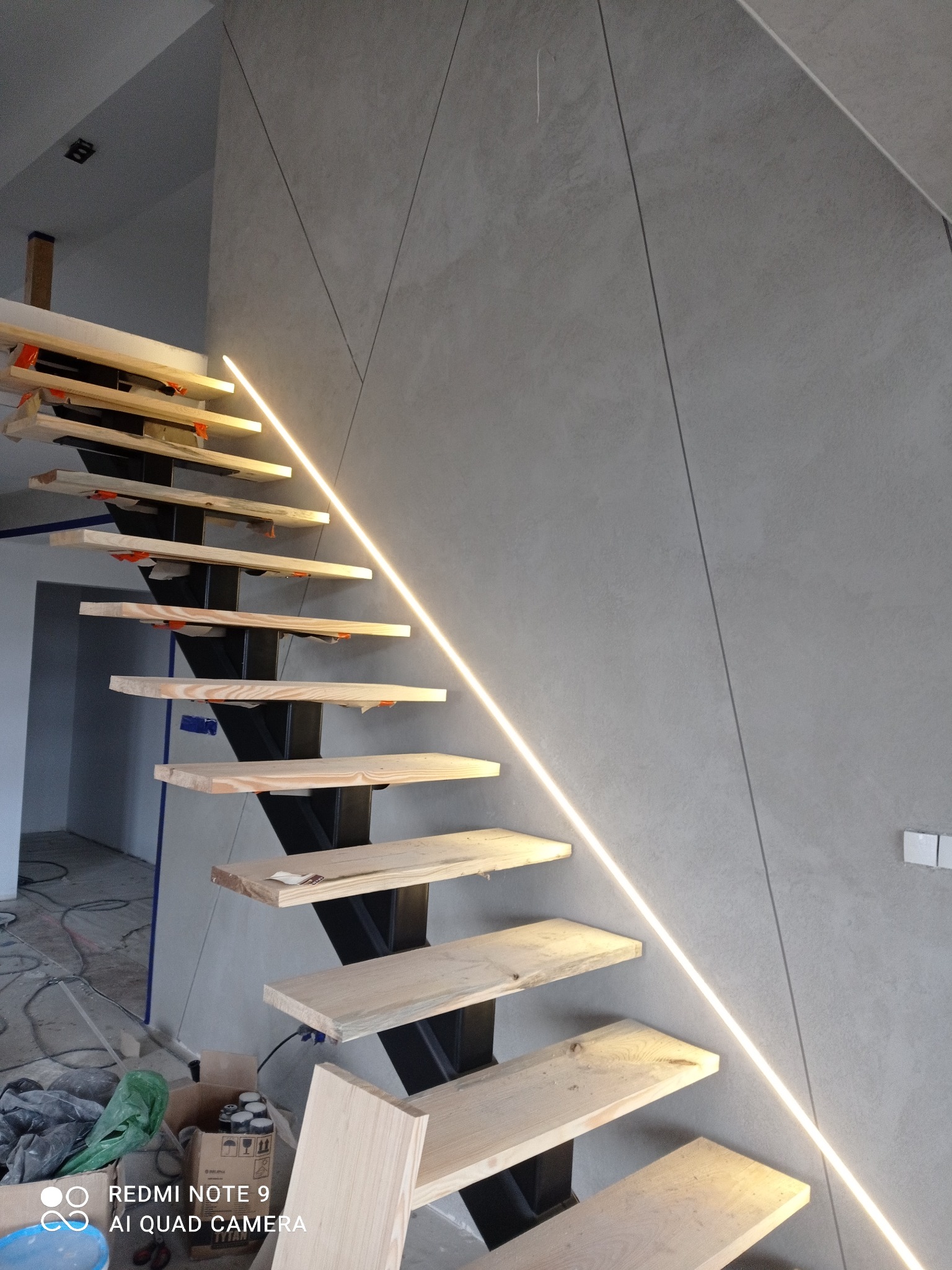 Surowe drewniane stopnie schodów na metalowej konstrukcji, podświetlone listwą LED na tle ściany z szarego betonu architektonicznego z widocznymi kablami i elementami wykończeniowymi w tle.