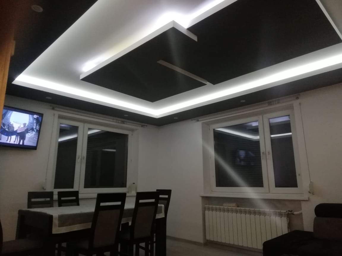 Nowoczesny salon z czarno-białym sufitem podwieszanym z oświetleniem LED, stół z krzesłami, okna i telewizor na ścianie.