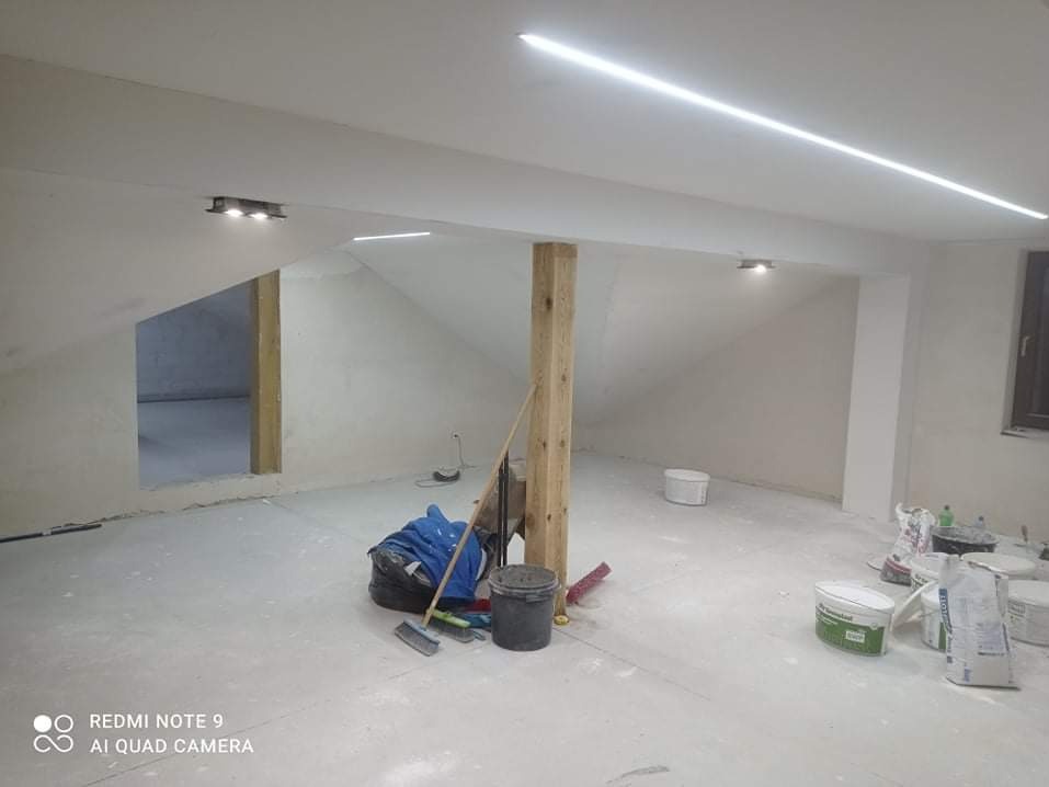 Poddasze w trakcie prac wykończeniowych, widoczne szpachlowane ściany, surowy drewniany słup, pędzle, wiadra z farbą i oświetlenie LED na suficie.