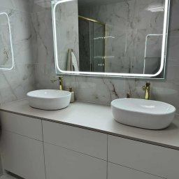 Latający Warsztat - Nowoczesna łazienka z dwoma białymi umywalkami nablatowymi, złotymi bateriami i podświetlanym lustrem na tle marmurowych płytek.