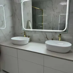 Nowoczesna łazienka z dwoma białymi umywalkami nablatowymi, złotymi bateriami i podświetlanym lustrem na tle marmurowych płytek.