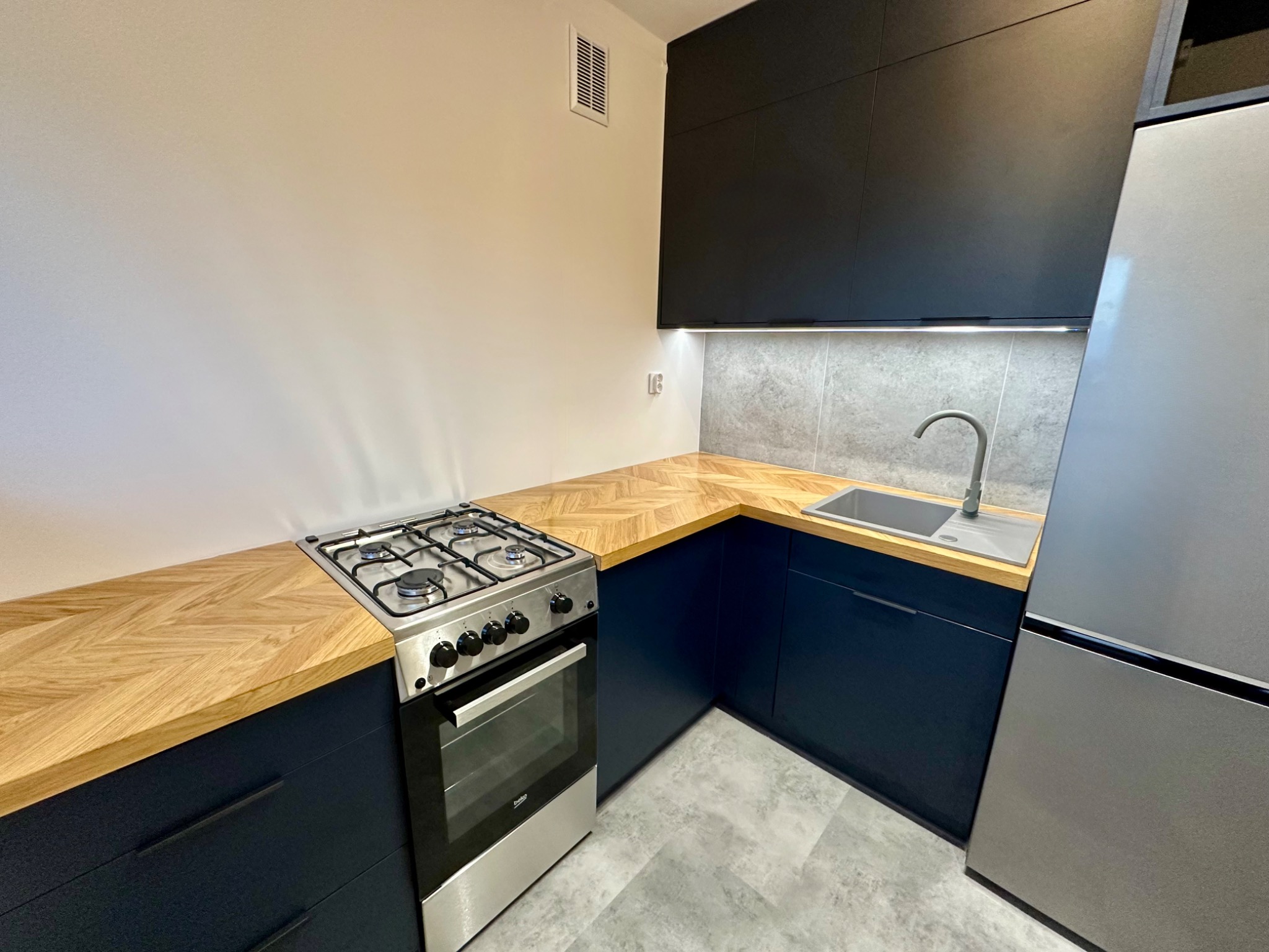 Niebieska zabudowa kuchenna z drewnianym blatem ułożonym w jodełkę, srebrny piekarnik i lodówka. Górne szafki ciemnoszare, podświetlane. Szare płytki na ścianie.