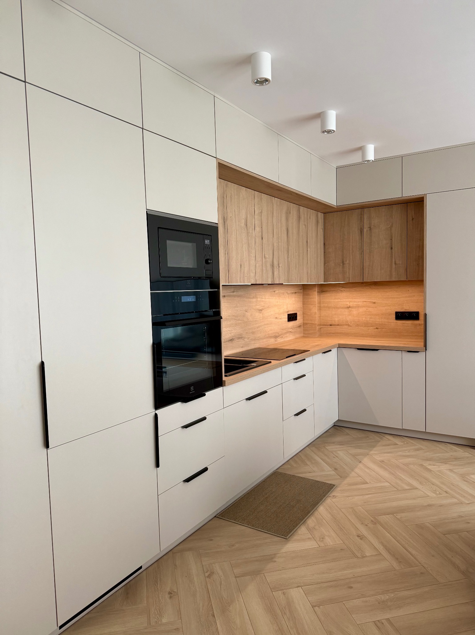 Biała kuchnia z drewnianymi akcentami: zabudowa po sufit, piekarnik i mikrofalówka w słupku, parkiet w jodełkę, minimalistyczne uchwyty, oświetlenie punktowe.