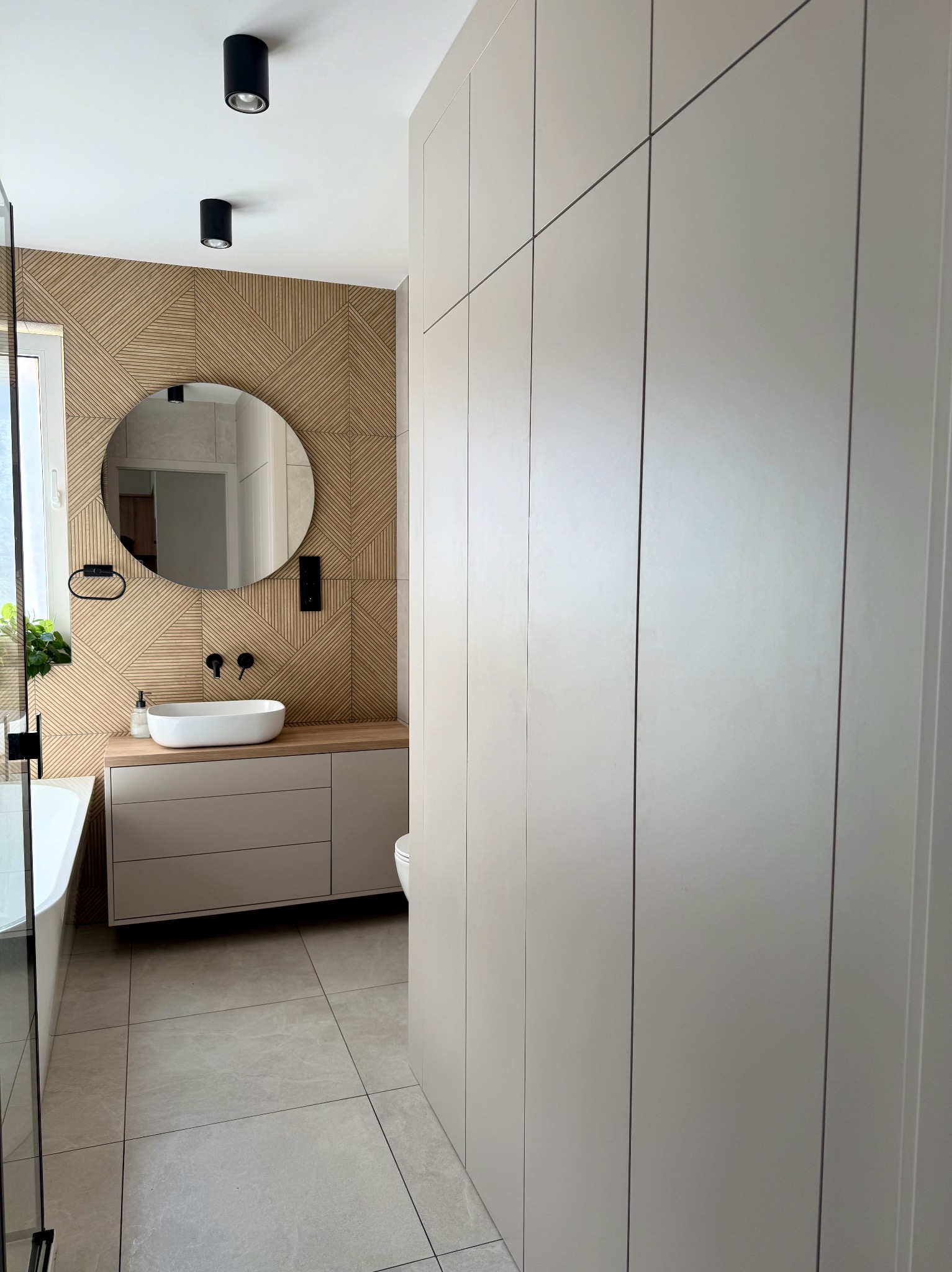 Nowoczesna łazienka z drewnianą ścianą za okrągłym lustrem, umywalką nablatową i beżowymi szafkami oraz wysoką zabudową meblową po prawej stronie.