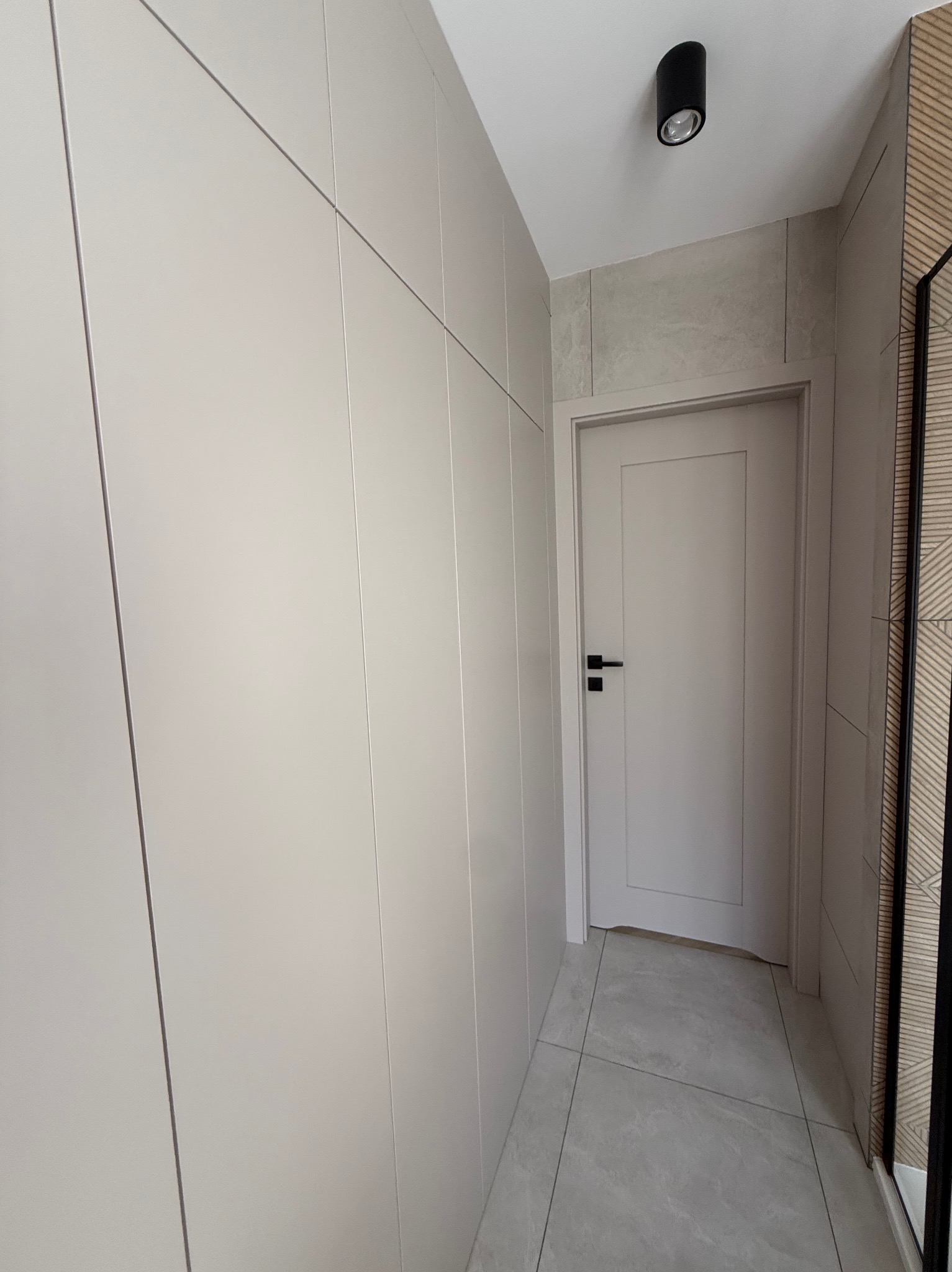 Jasna szafa wnękowa z gładkimi frontami, dopasowana do koloru drzwi we Wschowie. Minimalistyczny design, szare płytki podłogowe i sufitowe oświetlenie.