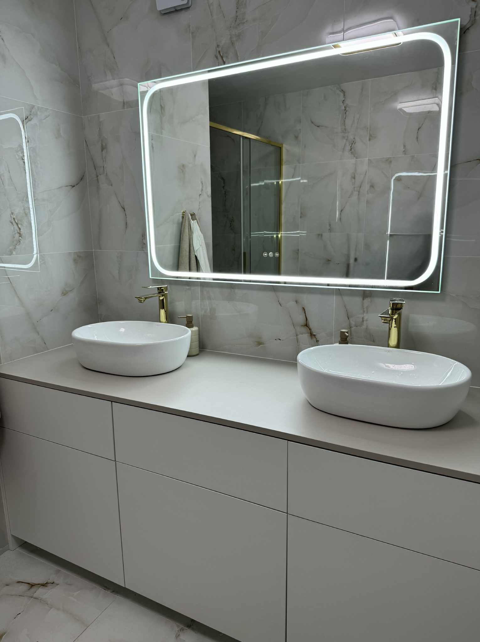 Nowoczesna łazienka z dwoma białymi umywalkami nablatowymi, złotymi bateriami i podświetlanym lustrem na tle marmurowych płytek.
