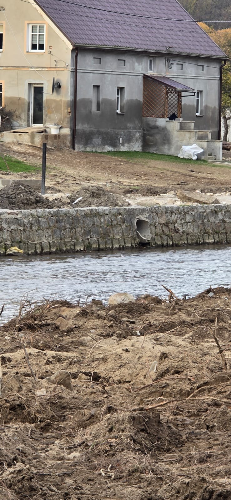 Brzeg rzeki umocniony kamiennym murem z betonową rurą odpływową, w tle dwa budynki mieszkalne po remoncie.