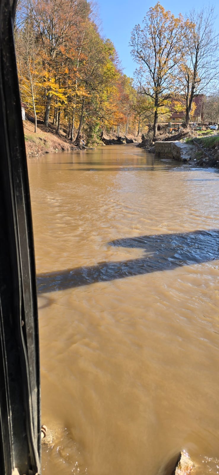 Mętna rzeka z brązową wodą płynąca przez obszar z drzewami w jesiennych kolorach, widziana z wnętrza kabiny maszyny budowlanej.