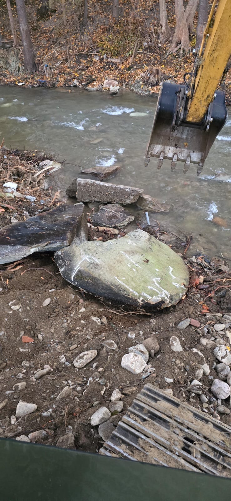 Żółta koparka umieszcza kamienie w korycie rzeki podczas prac regulacyjnych, widoczne zanieczyszczenia i brudna woda.