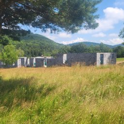 MĘŻYK-BUD MARCIN MĘŻYK - Niedokończony budynek z szarego bloczka na tle łąki i zalesionych wzgórz w słoneczny dzień, widoczny częściowo pod drzewem iglastym, z koparką w tle.