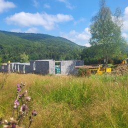 MĘŻYK-BUD MARCIN MĘŻYK - Wznoszone mury domu z betonu komórkowego na tle zalesionych wzgórz i błękitnego nieba z chmurami, na pierwszym planie wysoka trawa i kwiaty, obok budynku stoi żółta koparka.