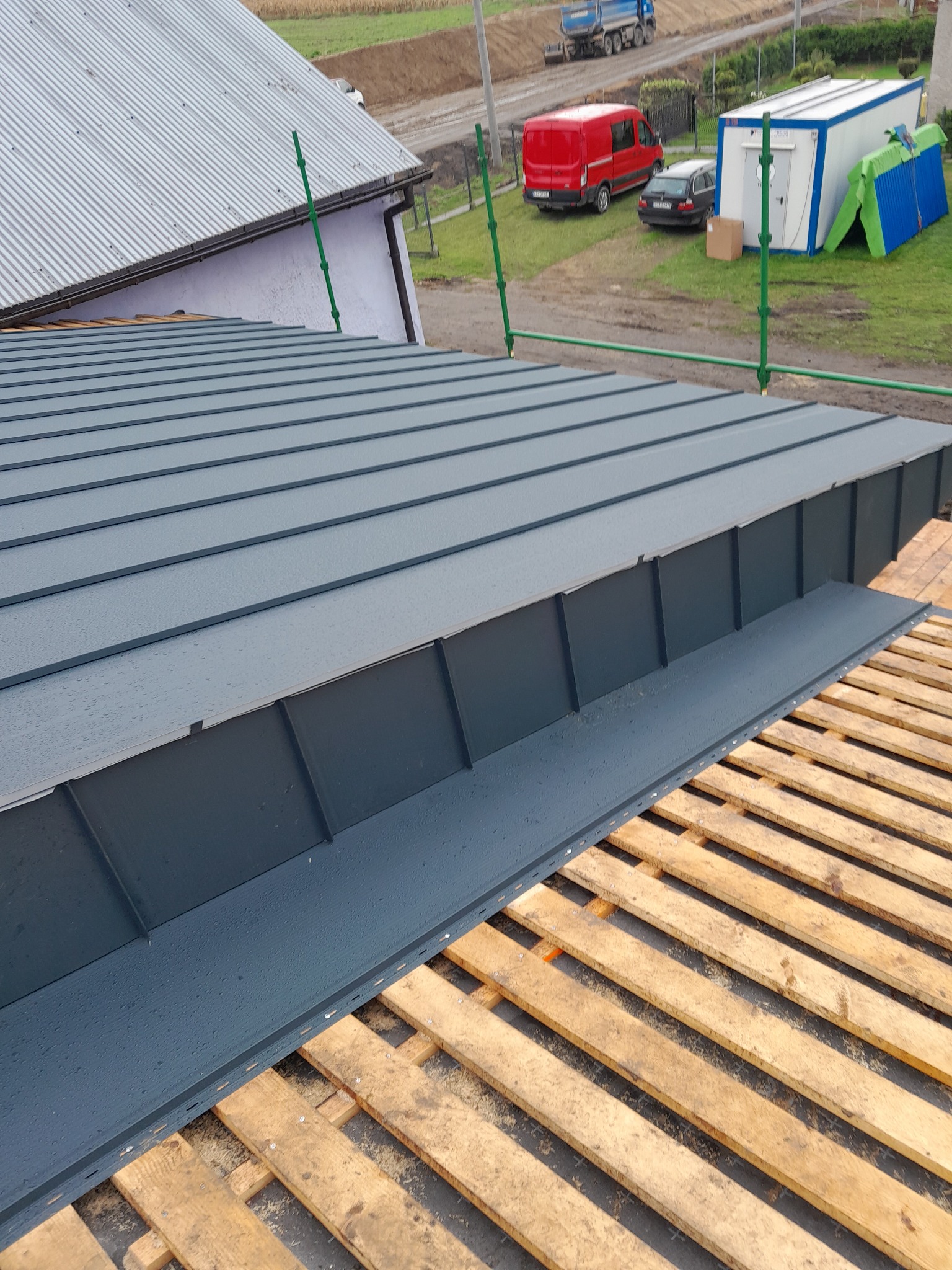 Montaż blachy na dachu, widoczne drewniane łaty i fragment nowego orynnowania w kolorze grafitowym, w tle budynek gospodarczy, samochody i kontener biurowy.
