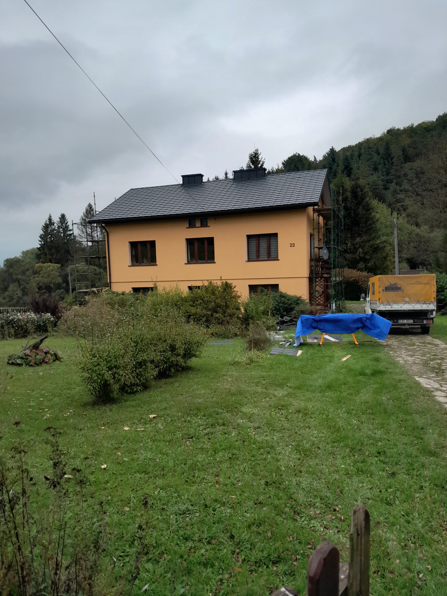 Żółty dom jednorodzinny z nowym szarym dachem, otoczony rusztowaniami, z niebieską plandeką i samochodem dostawczym w tle, na tle zalesionego wzgórza w pochmurny dzień.