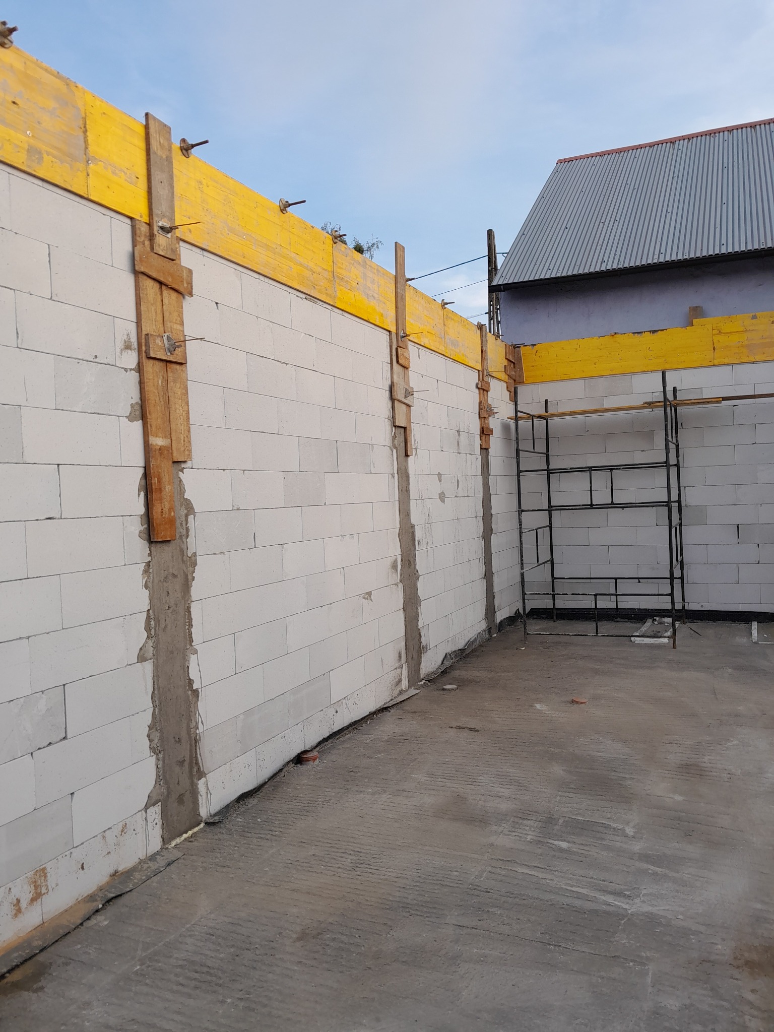 Wznoszenie ścian z białych bloczków z widocznym szalunkiem i podporami drewnianymi, na przygotowanym fundamencie, z rusztowaniem w tle i fragmentem budynku sąsiedniego.