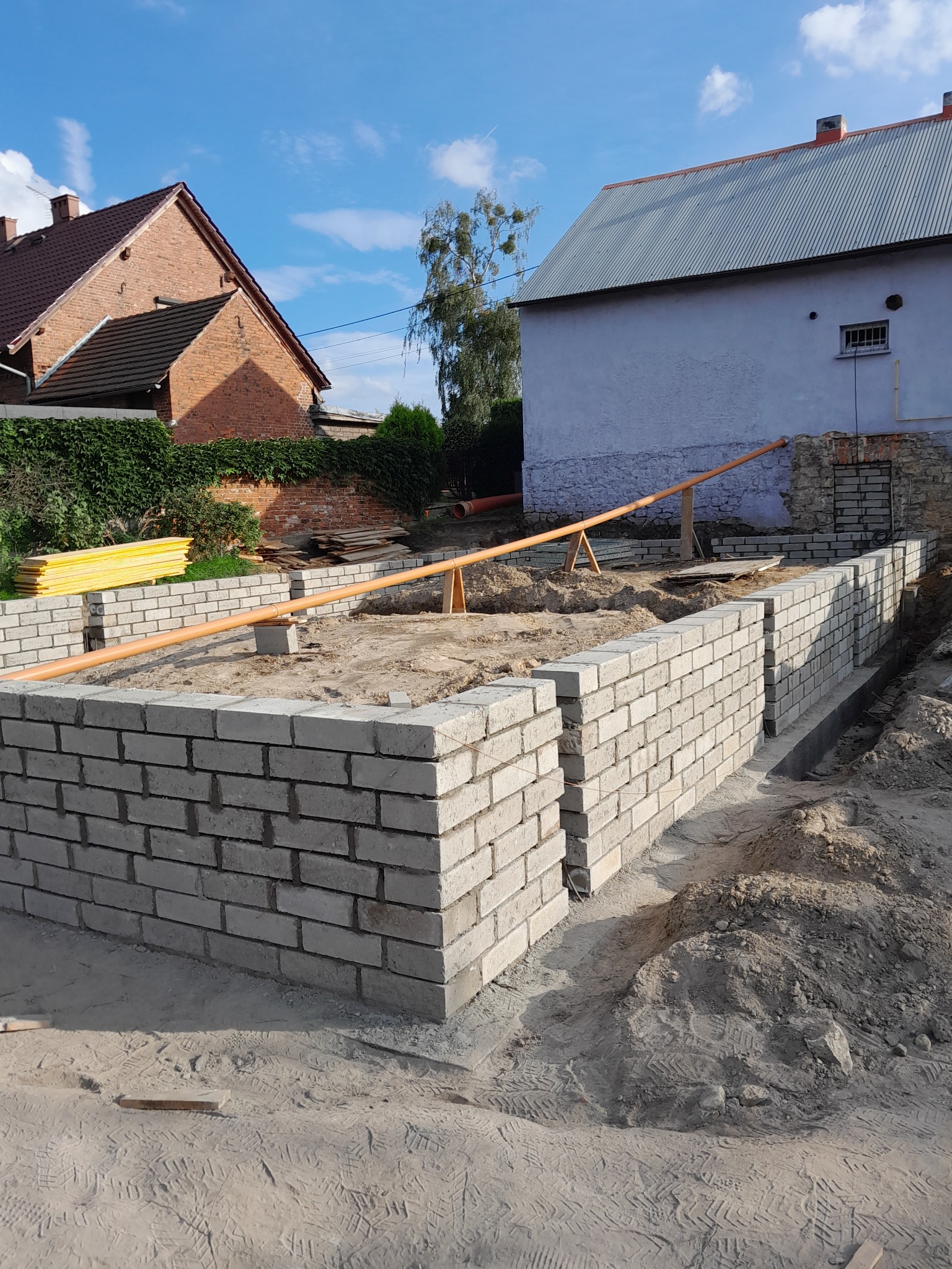 Wznoszenie fundamentów z białej cegły silikatowej na budowie domu, widoczne rury pomiarowe, w tle budynki mieszkalne i błękitne niebo.
