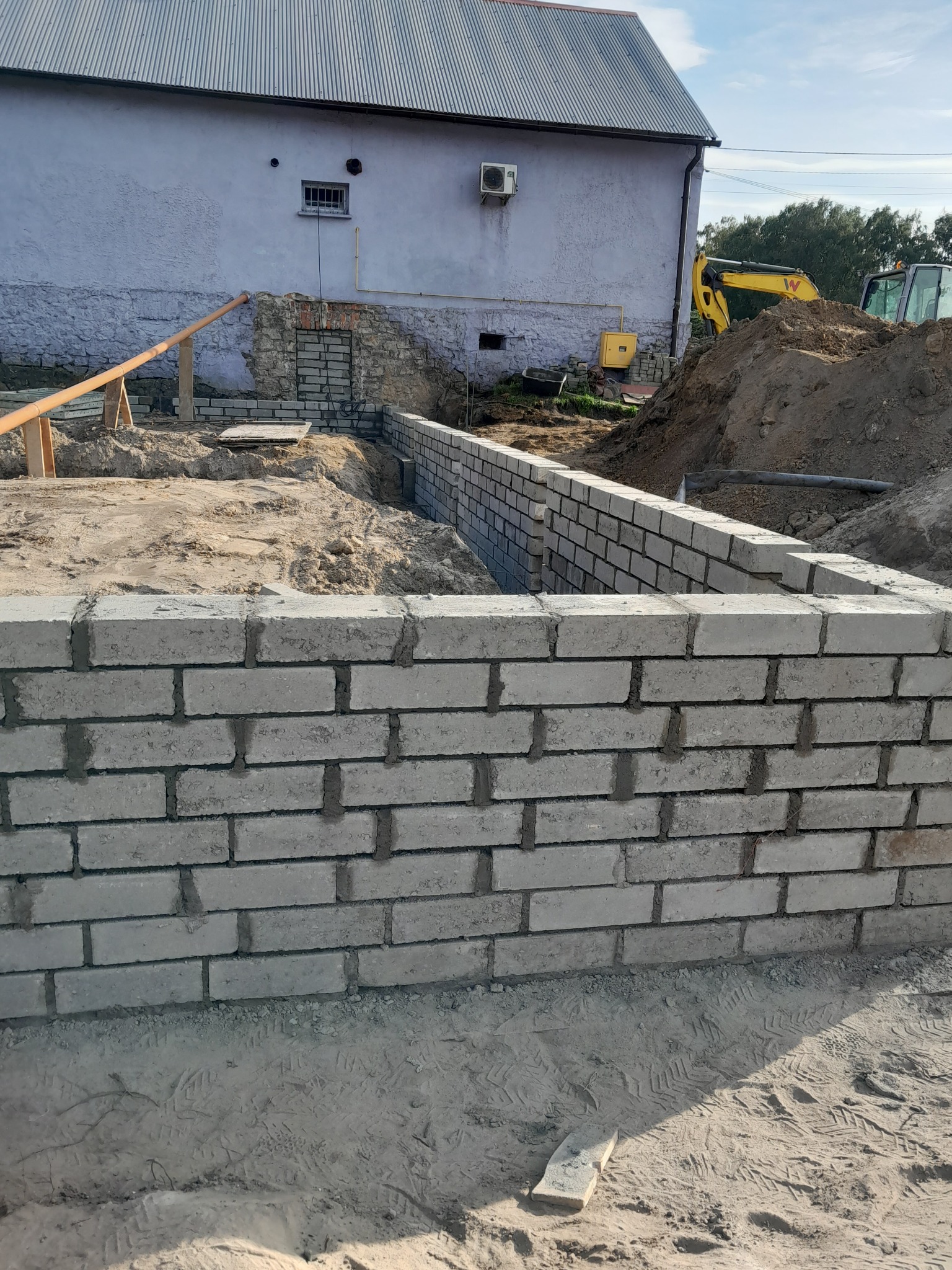 Wznoszenie fundamentów z betonowych bloczków na placu budowy, widoczny wykop i koparka w tle.