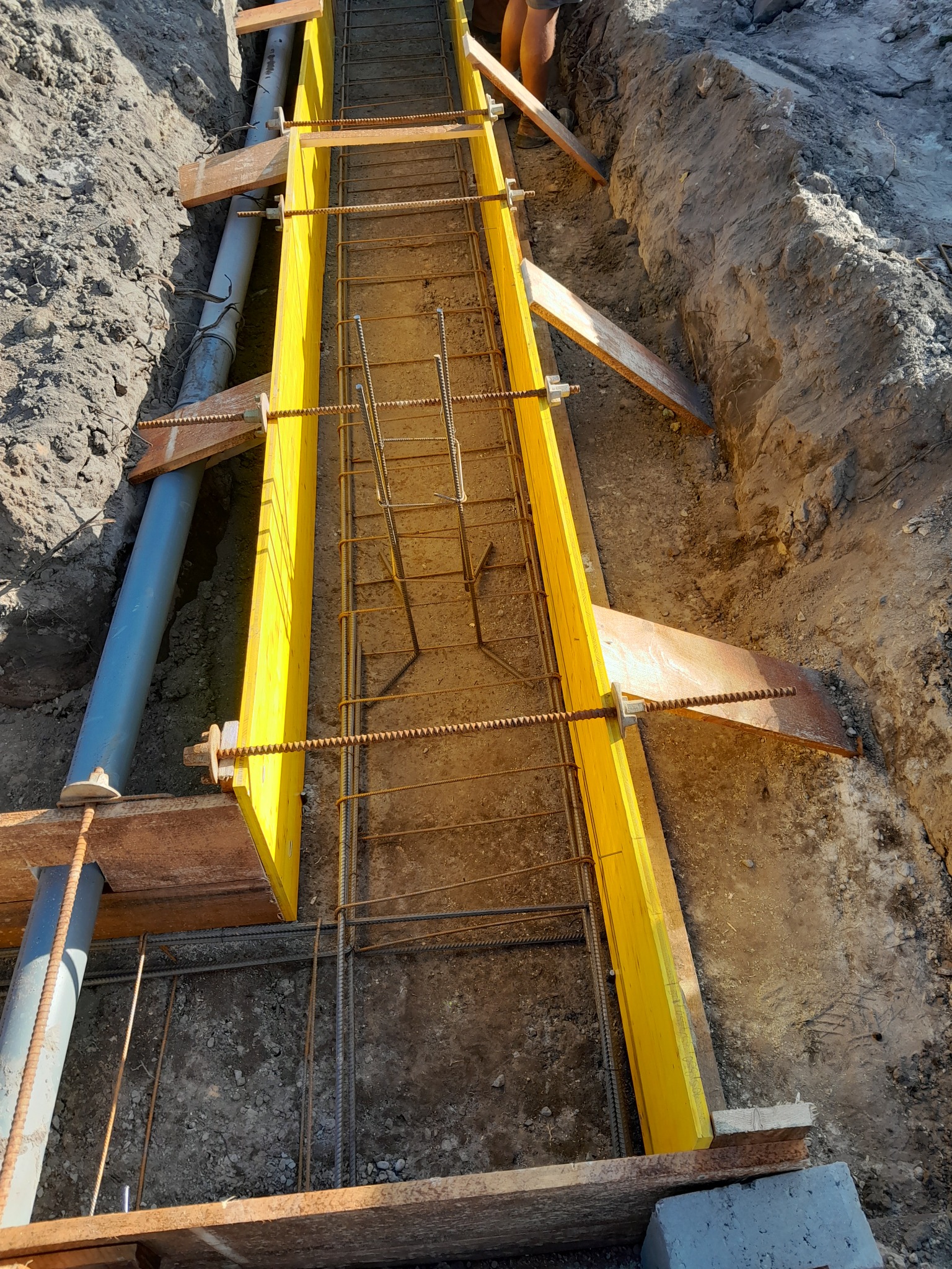 Przygotowanie szalunku i zbrojenia pod fundament w Grybowie, widoczny wykop, żółte deski szalunkowe, stalowe pręty zbrojeniowe oraz rura kanalizacyjna.