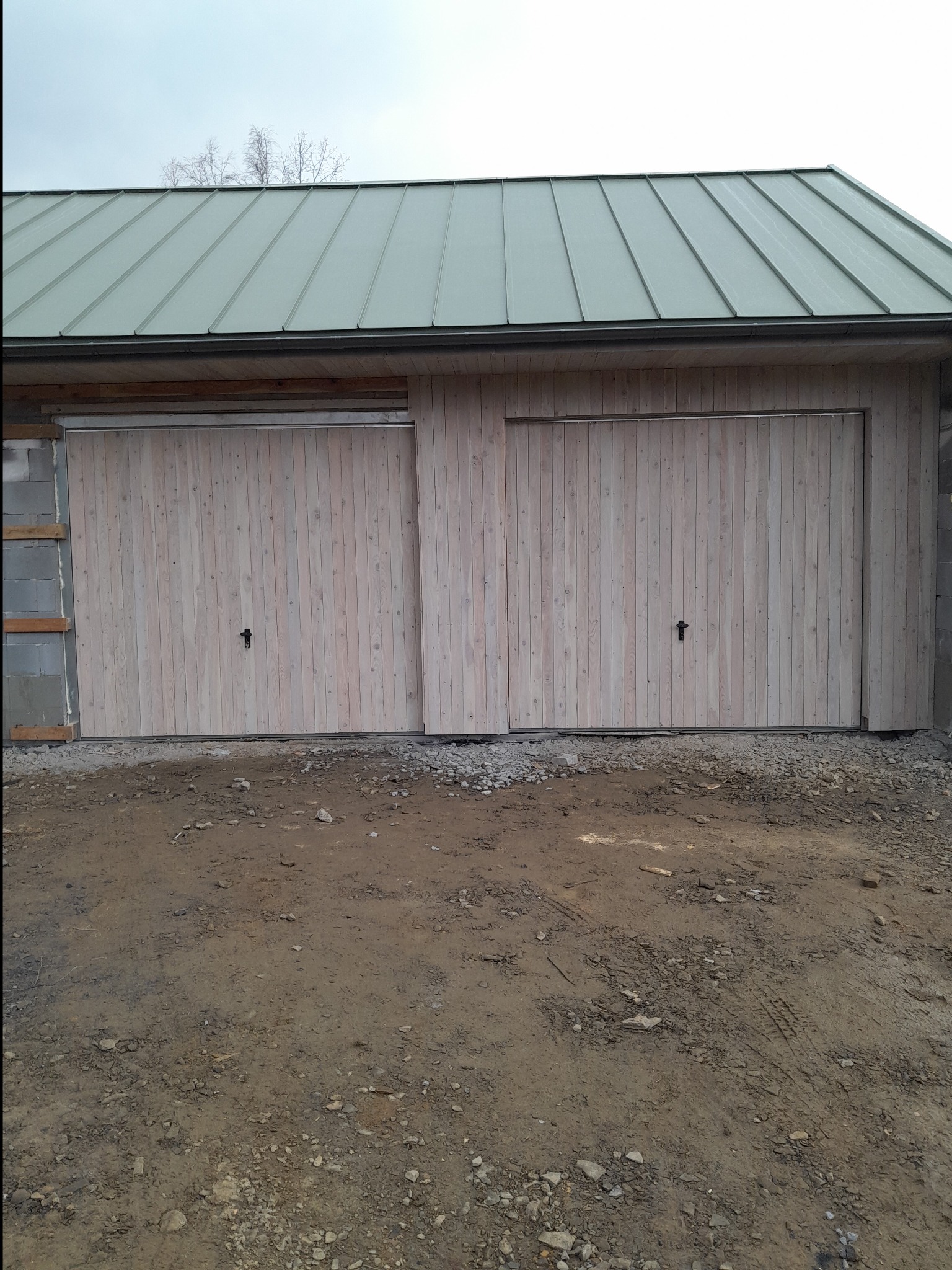 Budynek w stanie surowym zamkniętym z drewnianymi drzwiami garażowymi i zielonym dachem z blachy, częściowo ocieplony, widok na teren budowy.