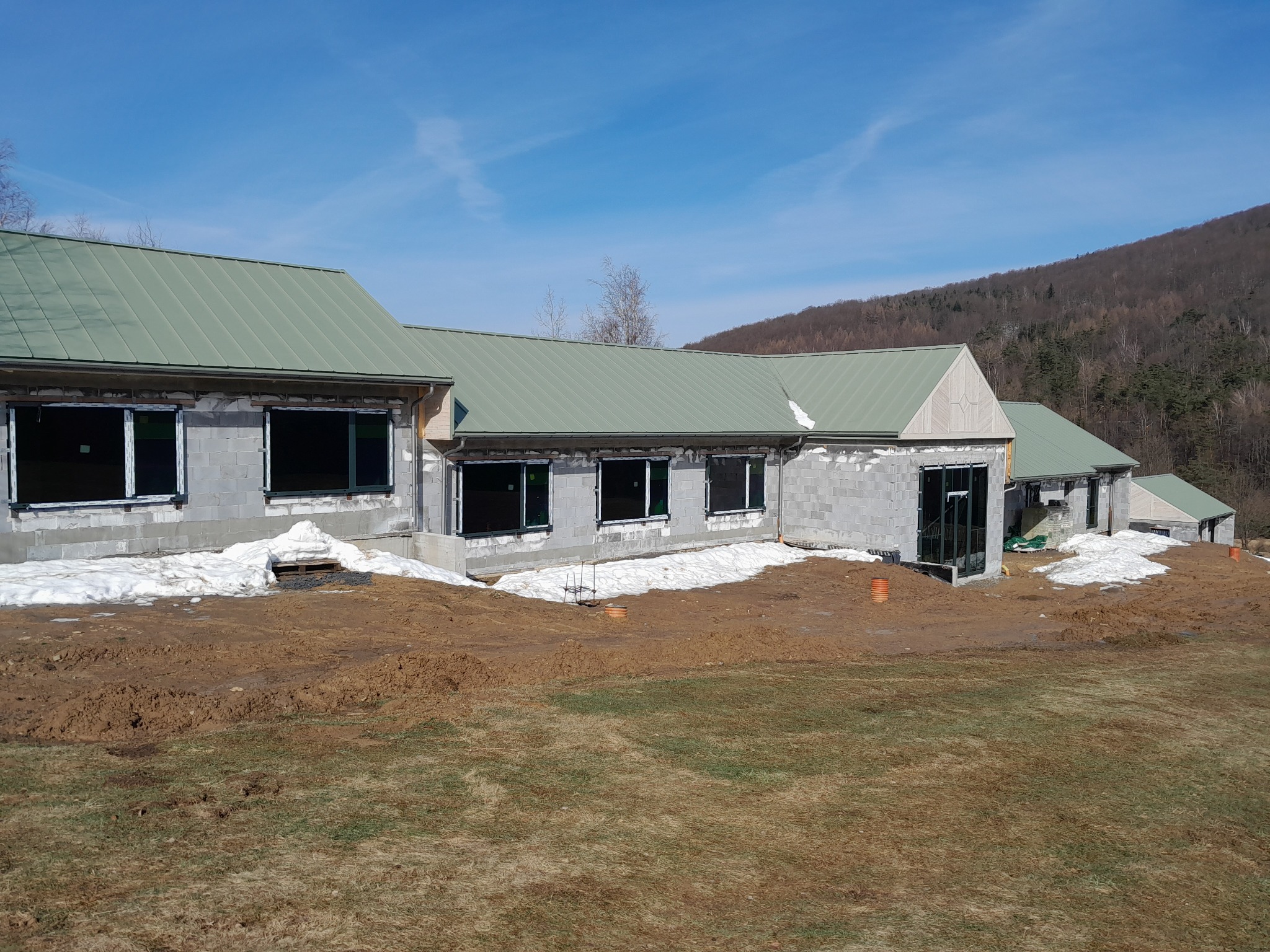 Wznoszony budynek z szarego bloczka, z zielonym dachem, w stanie surowym otwartym, z widocznymi oknami i drzwiami, na tle zalesionego wzgórza i błękitnego nieba, z pozostałościami śniegu przy...